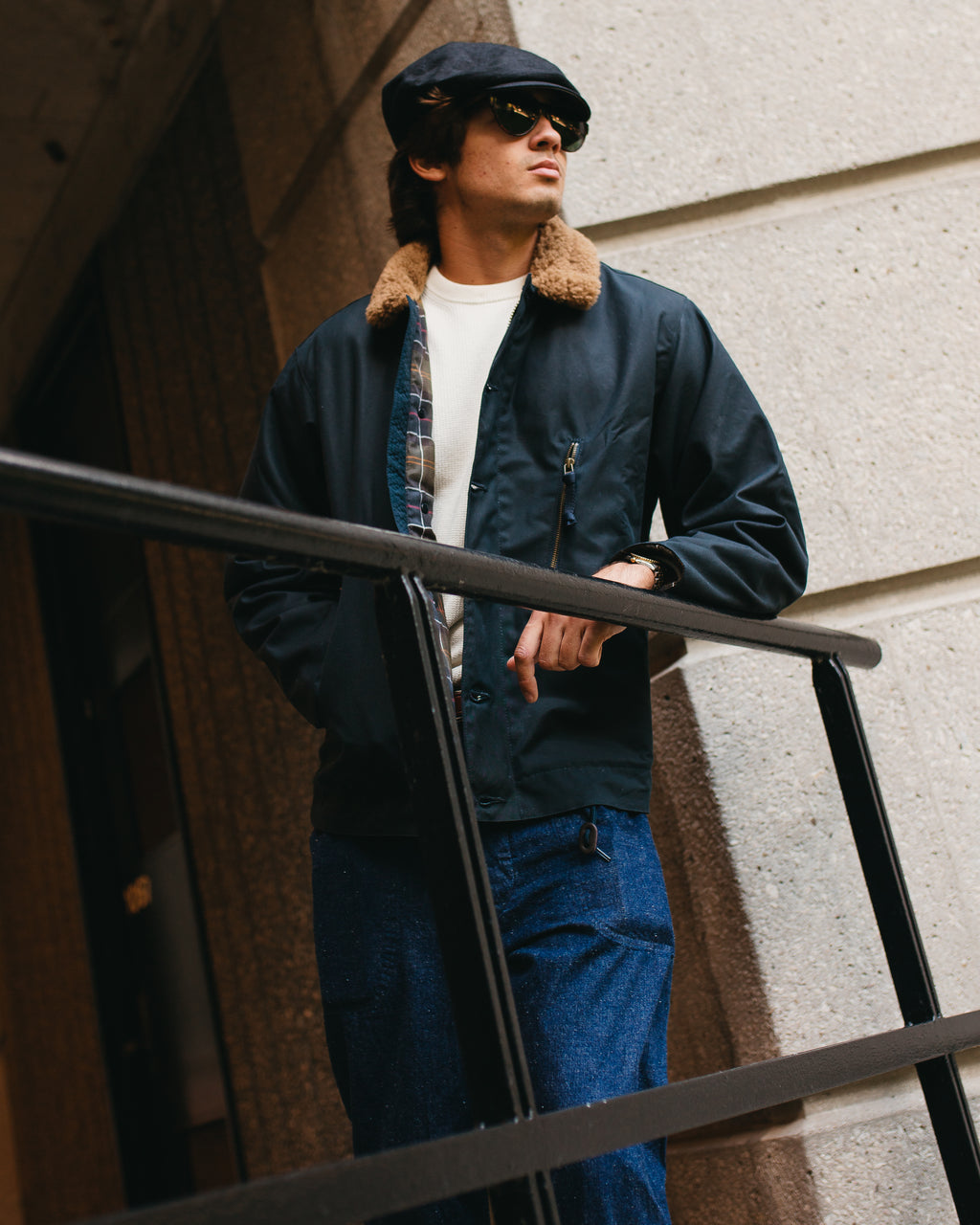 A man wearing a navy blue waxed cotton bomber jacket with a brown shearling collar, blue jeans, a white t-shirt, and a black cap, leaning on a black metal railing against a stone wall.