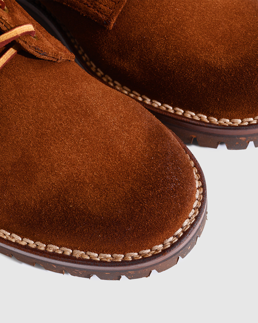 Close-up of a brown suede boot with tan contrast stitching and a dark brown sole, shown against a light gray background.