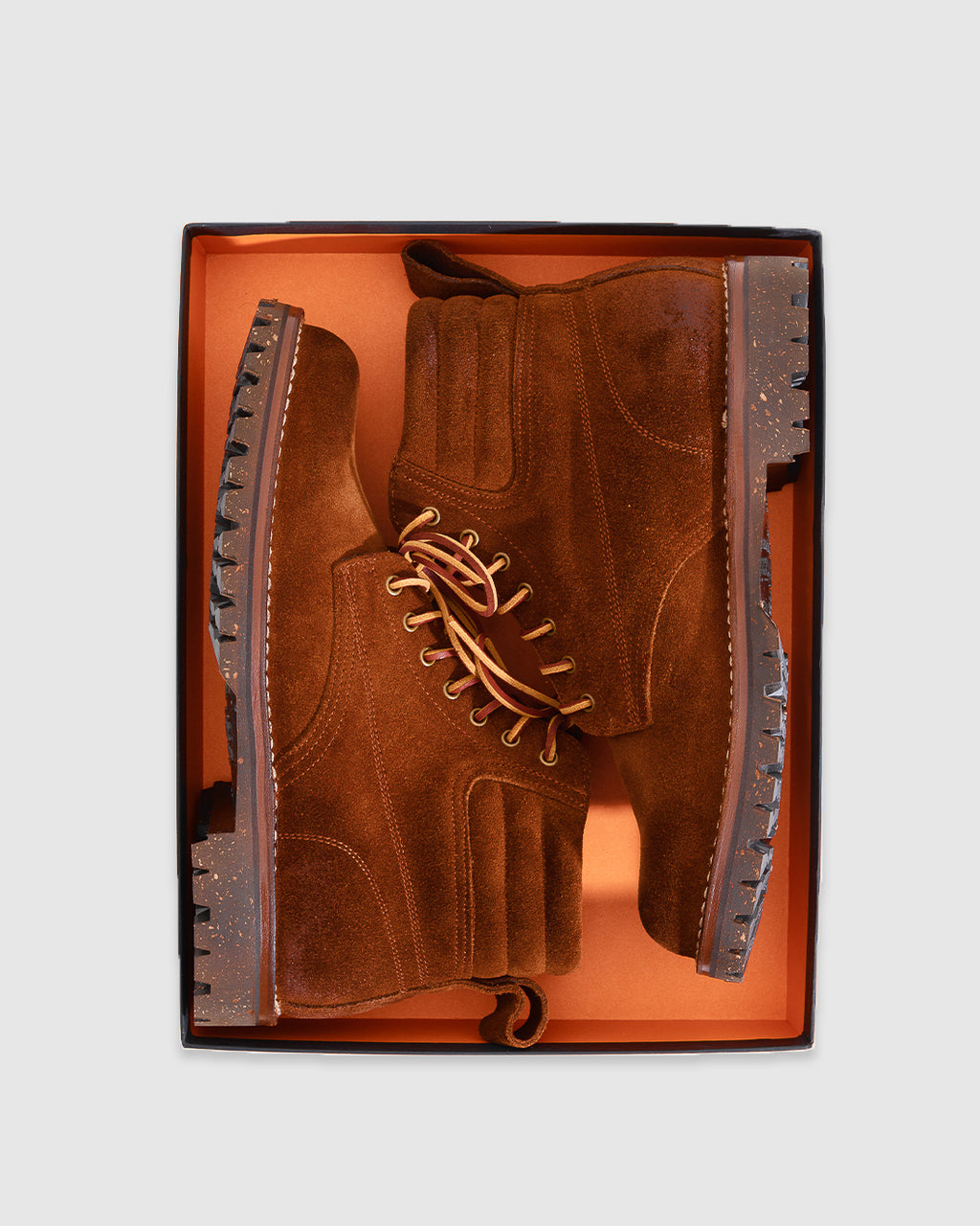A pair of brown suede lace-up boots displayed inside an open orange shoebox against a light gray background.
